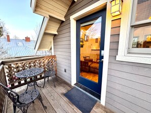 Une terrasse en bois avec deux tables et des chaises en métal, une porte bleue donnant sur un espace intérieur, et une vue sur les maisons voisines.