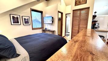 Une chambre avec un grand lit, une table en bois, un ventilateur de plafond et un téléviseur fixé au mur.