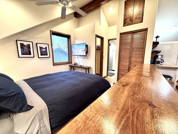 Une chambre avec un grand lit, une table en bois, un ventilateur de plafond et un téléviseur fixé au mur.