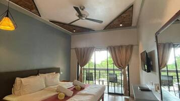 A bedroom with a bed, ceiling fan, large windows with curtains, and a TV mounted on the wall.