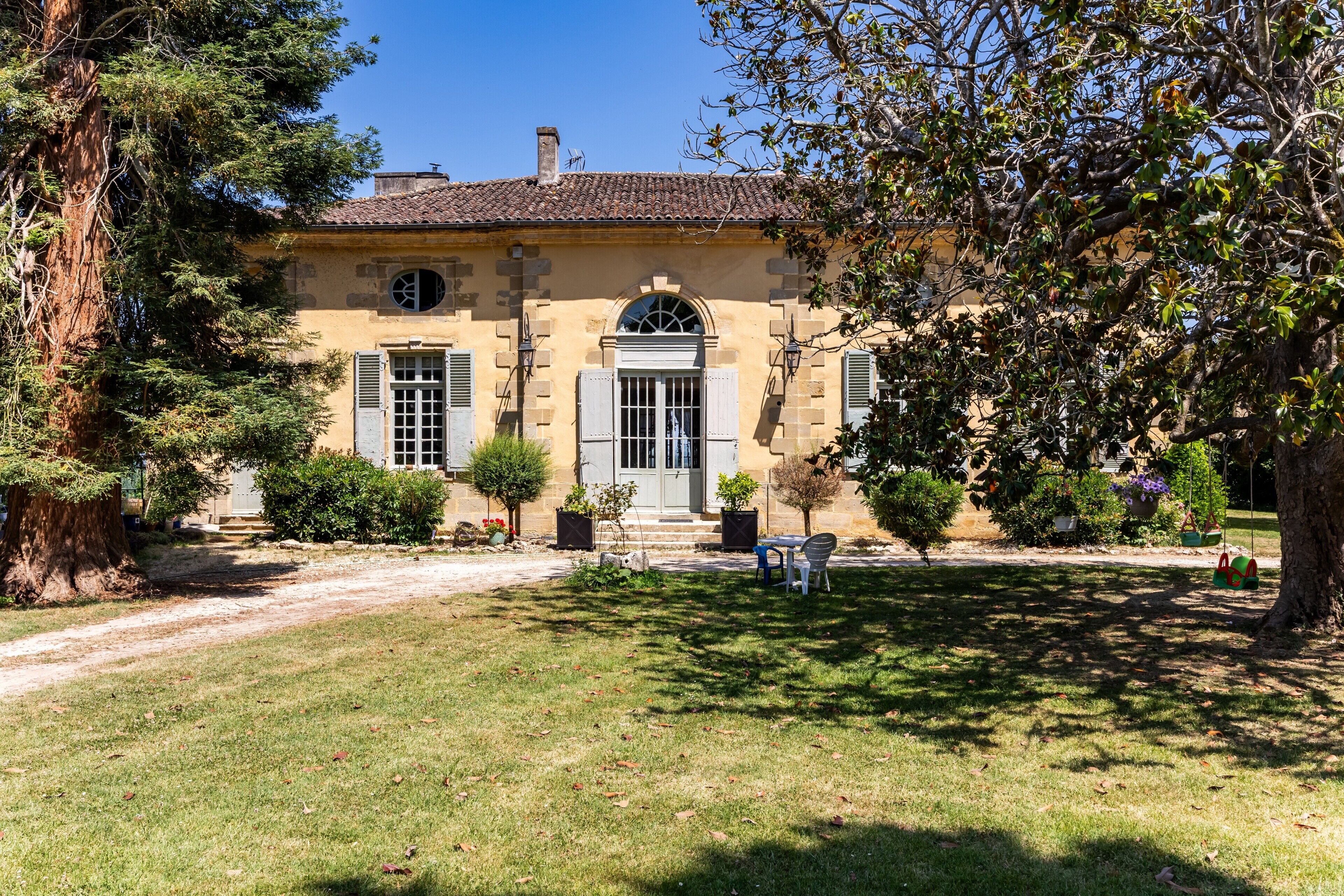 A traditional stone building with a tiled roof, a central entrance, and a surrounding garden with trees and a pathway.
