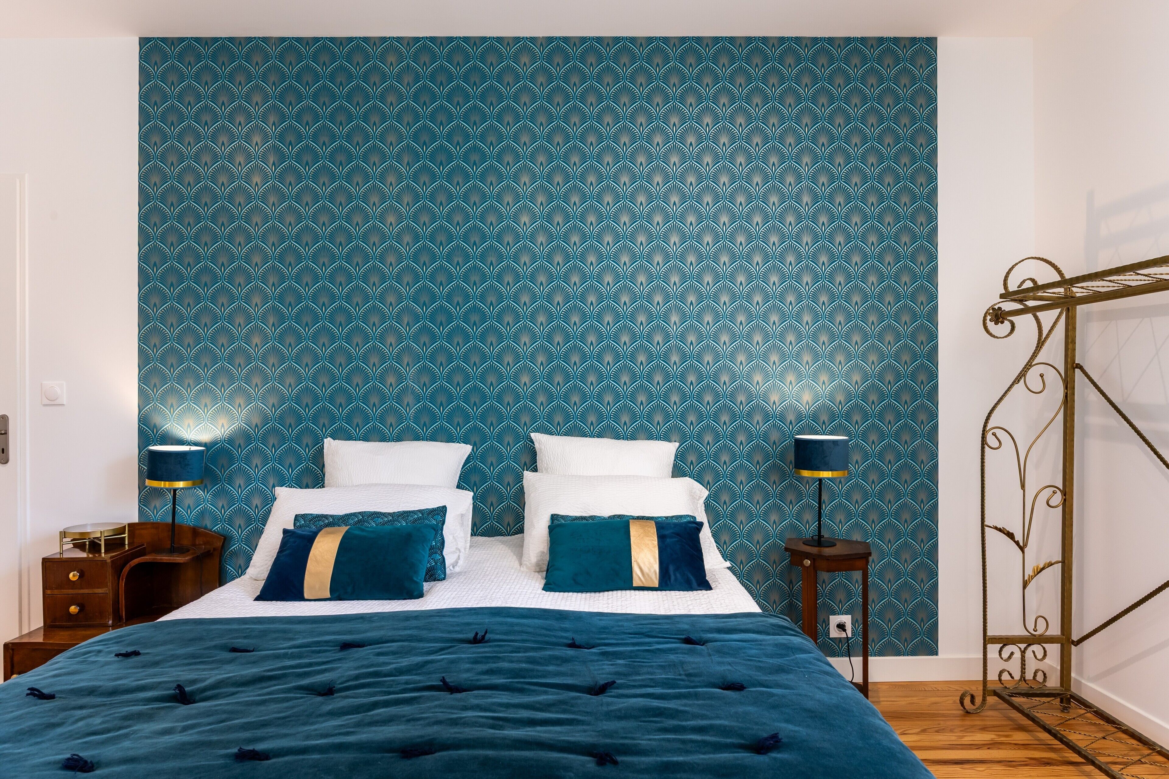 Une chambre à coucher avec un papier peint à motifs, un lit avec de nombreux coussins, deux tables de chevet et une lampe sur pied.
