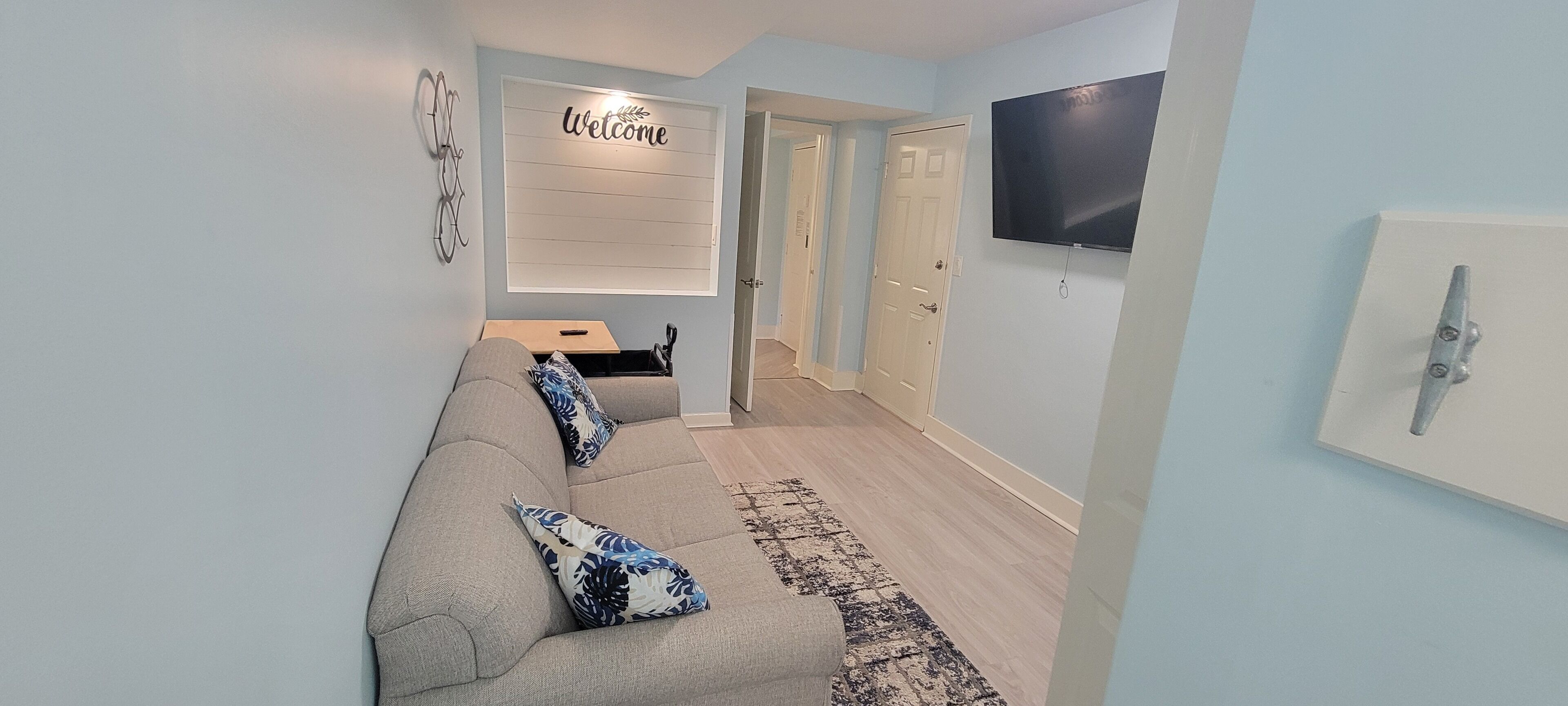 Ein modernes Wohnzimmer mit einem grauen Sofa, gemusterten Kissen, einem wandmontierten Fernseher und einem 'Welcome'-Schild an der Wand.