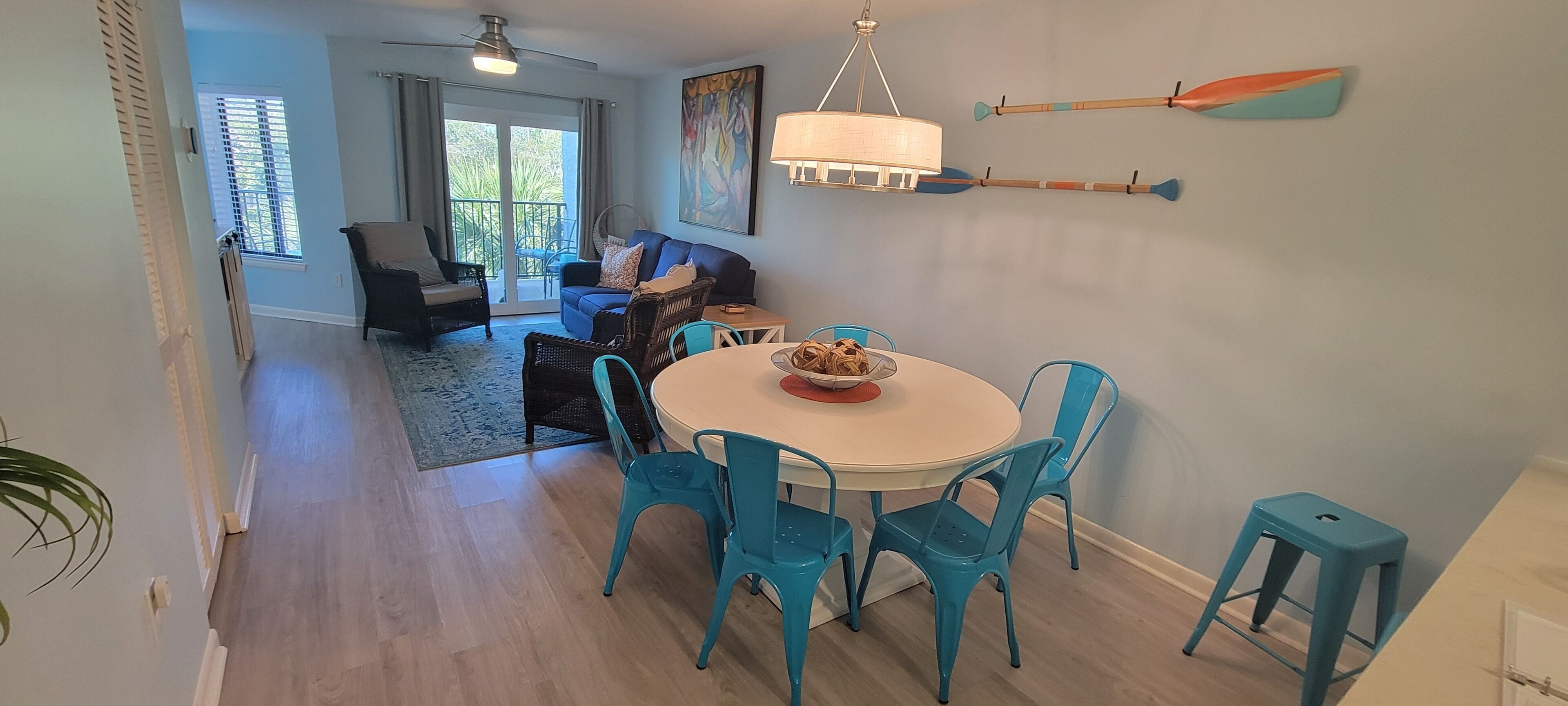 A dining area with a round table, blue chairs, and a bowl of fruit. There is a sofa and chairs in the background, and a window with blinds.