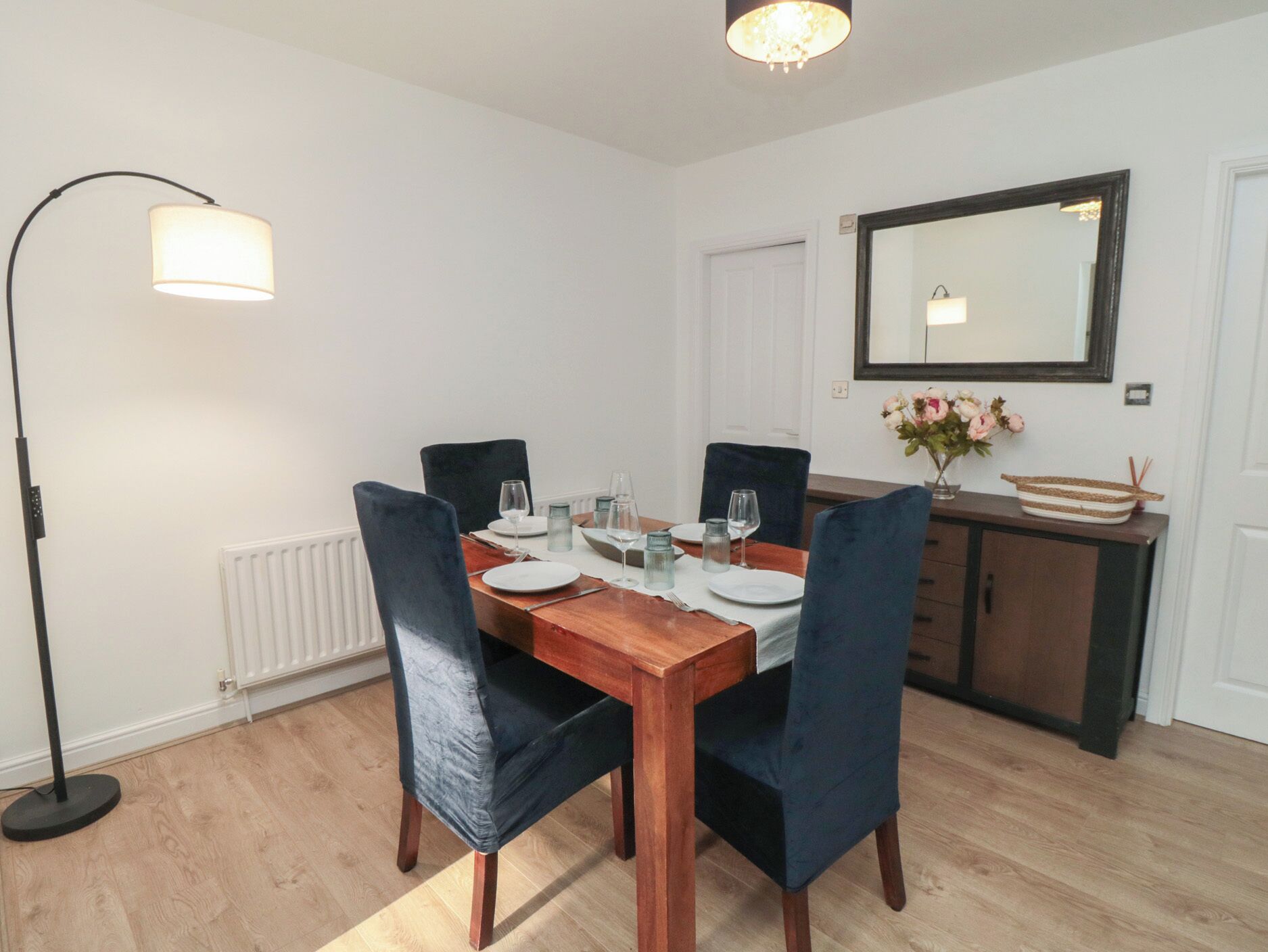 A dining area with a wooden table set for four, flanked by two blue chairs. A floor lamp is positioned to the left, and a mirror hangs on the wall above the table.