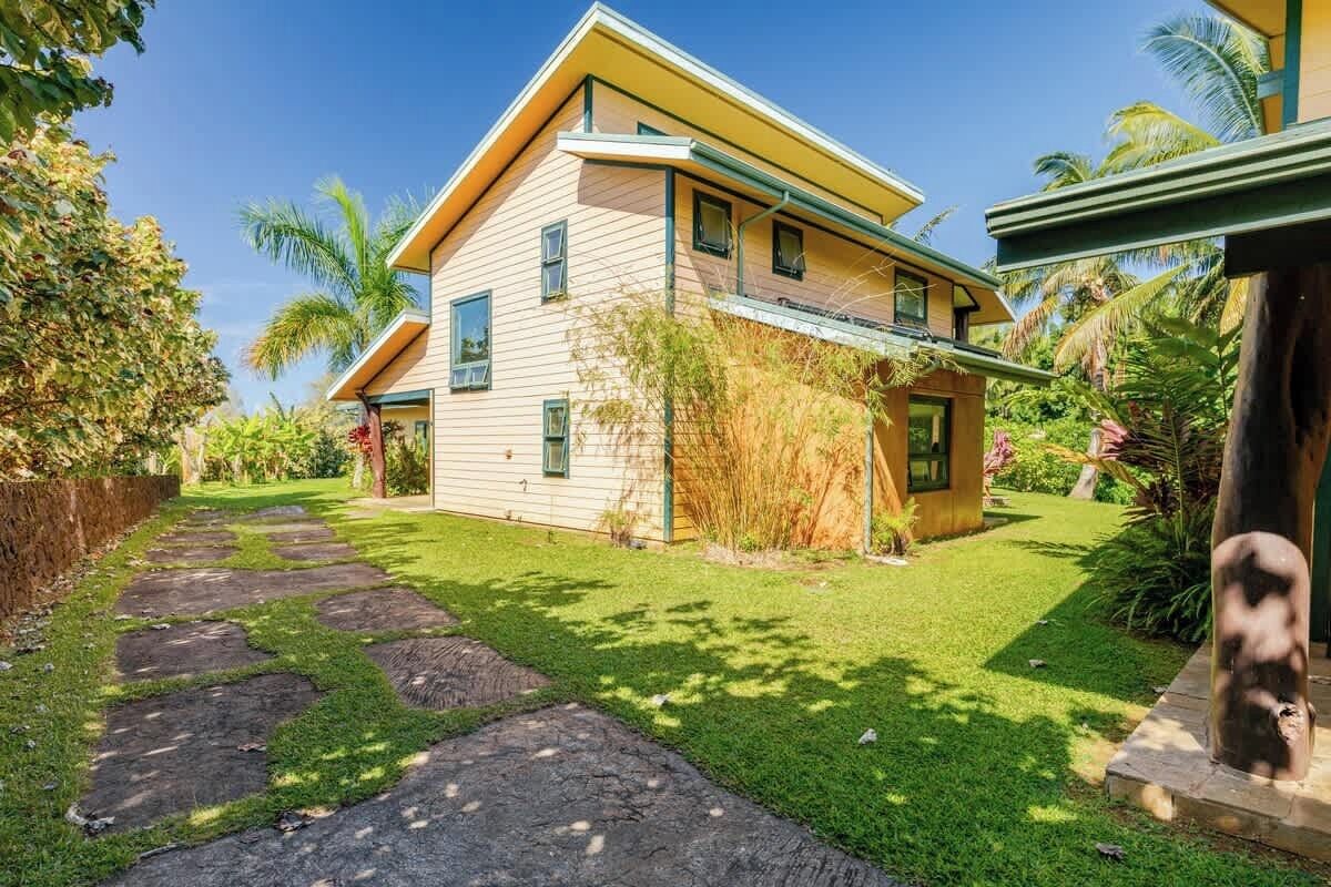 Une maison à deux étages, à la façade jaune, avec un chemin de pierre menant à la porte d’entrée et un jardin luxuriant agrémenté de palmiers et de plantes tropicales.