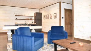 A room with a pool table, two blue chairs, a wooden coffee table, and a kitchen area with shelves and framed pictures. - Modern Getaway: Hot Tub, Pool Table, Arcade & Fire Pit (Blue Ridge)