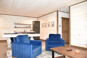 Ein Zimmer mit Pooltisch, zwei blauen Stühlen, einem Holztisch und einer Küchenzeile mit Regalen und gerahmten Bildern.