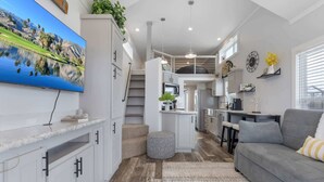 Un interno moderno con cucina, sala da pranzo e soggiorno, caratterizzato da mobili bianchi, un divano grigio e un grande televisore che mostra un paesaggio panoramico.
