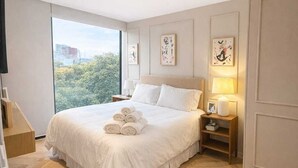 Hotellihuone, jossa on suuri sänky, yöpöytä, lamppuvarustettu sivupöytä ja suuri ikkuna, josta avautuu näkymä kaupunkiin.