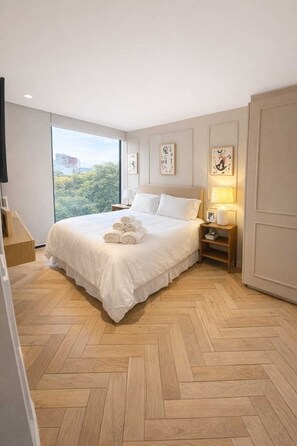 A hotel room with a large bed, a nightstand, a bedside table with a lamp, and a view of the city through a large window. - Casa Gama Colibri (Ciudad de México)
