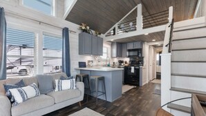 A spacious living area with a sofa, a kitchen with grey cabinets, and a loft bedroom with a wooden ceiling. - Little Blue Haus with Hot Tub! (Fredericksburg)