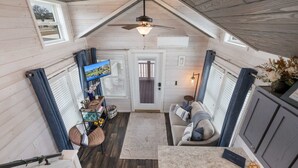 A loft-style room with a ceiling fan, a television, a dining area with a table and chairs, and a view of the outdoors. - Little Blue Haus with Hot Tub! (Fredericksburg)