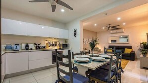 Una cocina moderna con armarios blancos, un comedor con mesa dispuesta para cuatro personas y una sala de estar con sofá y televisor.