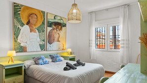 Ett sovrum med en säng, två sängbord, en hängande lampa, ett fönster med galler och konstverk på väggen.