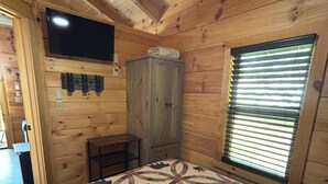 Ein Zimmer in einer Holzhütte mit einem Bett, einem an der Wand befestigten Fernseher, einem Fenster mit Jalousien und einem Deckenventilator.