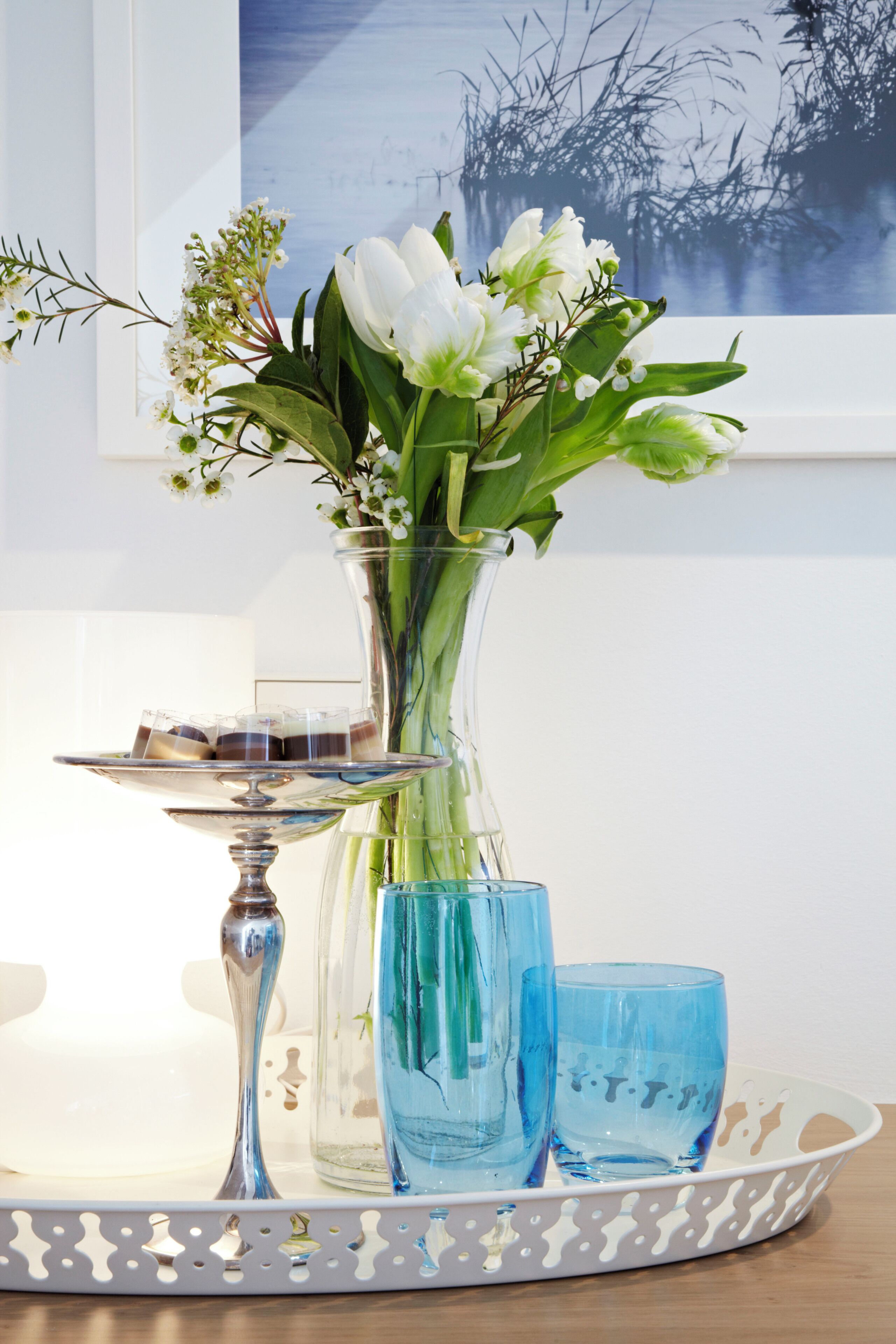 A clear glass vase with flowers and a tray with two blue glasses on a white table.