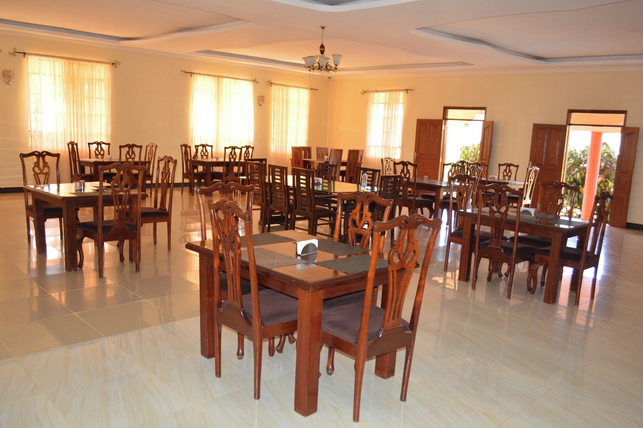 A spacious dining area with multiple wooden tables and chairs, large windows with curtains, and a chandelier.