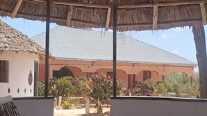 A covered outdoor seating area with a thatched roof, a view of a building with a metal roof, and a tiled floor. - Hoyjiifka Holale Resort (Moyale)