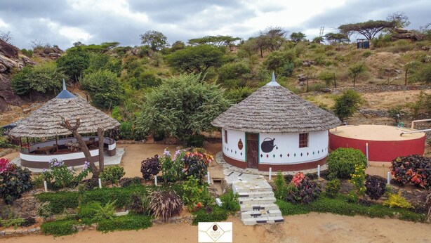 A rural setting with two thatched-roof huts, one with a circular design and another with a rectangular layout, surrounded by lush vegetation and a dirt path leading to a red structure. - Hoyjiifka Holale Resort (Moyale)