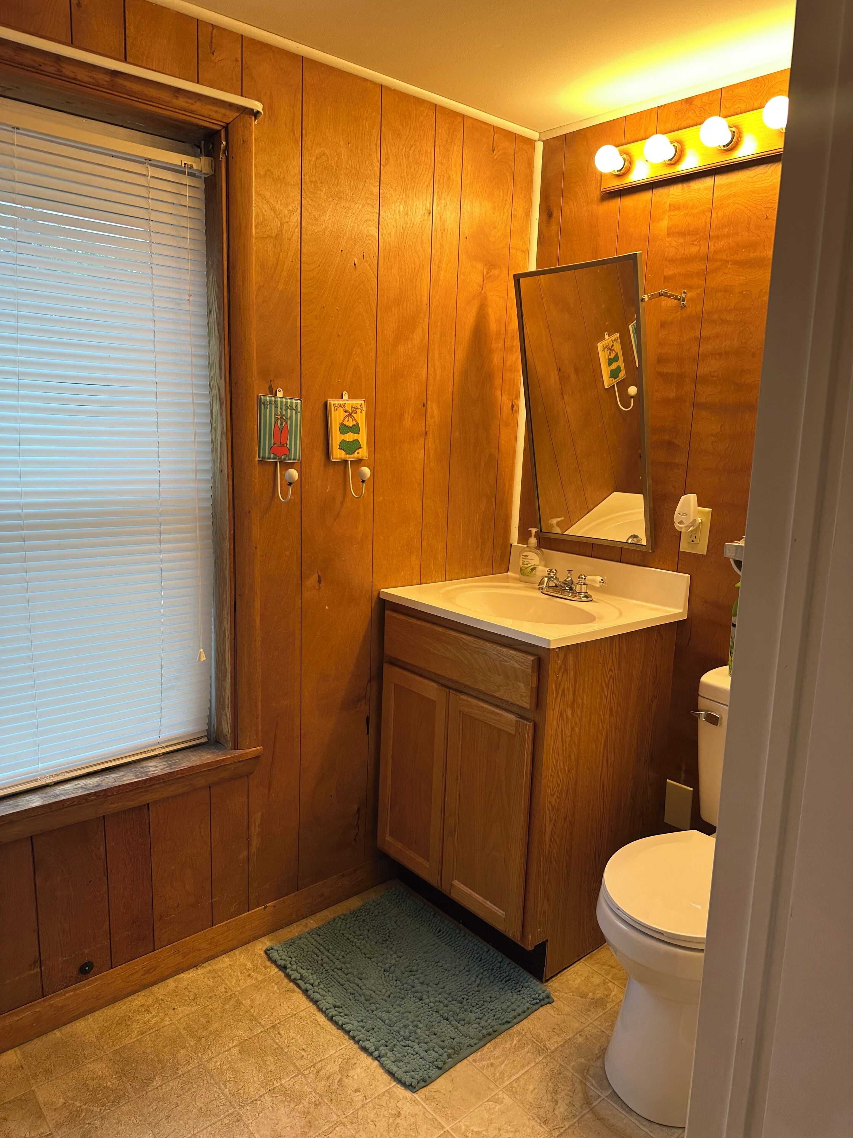A bathroom with wooden walls, a sink, a toilet, and a window with blinds.