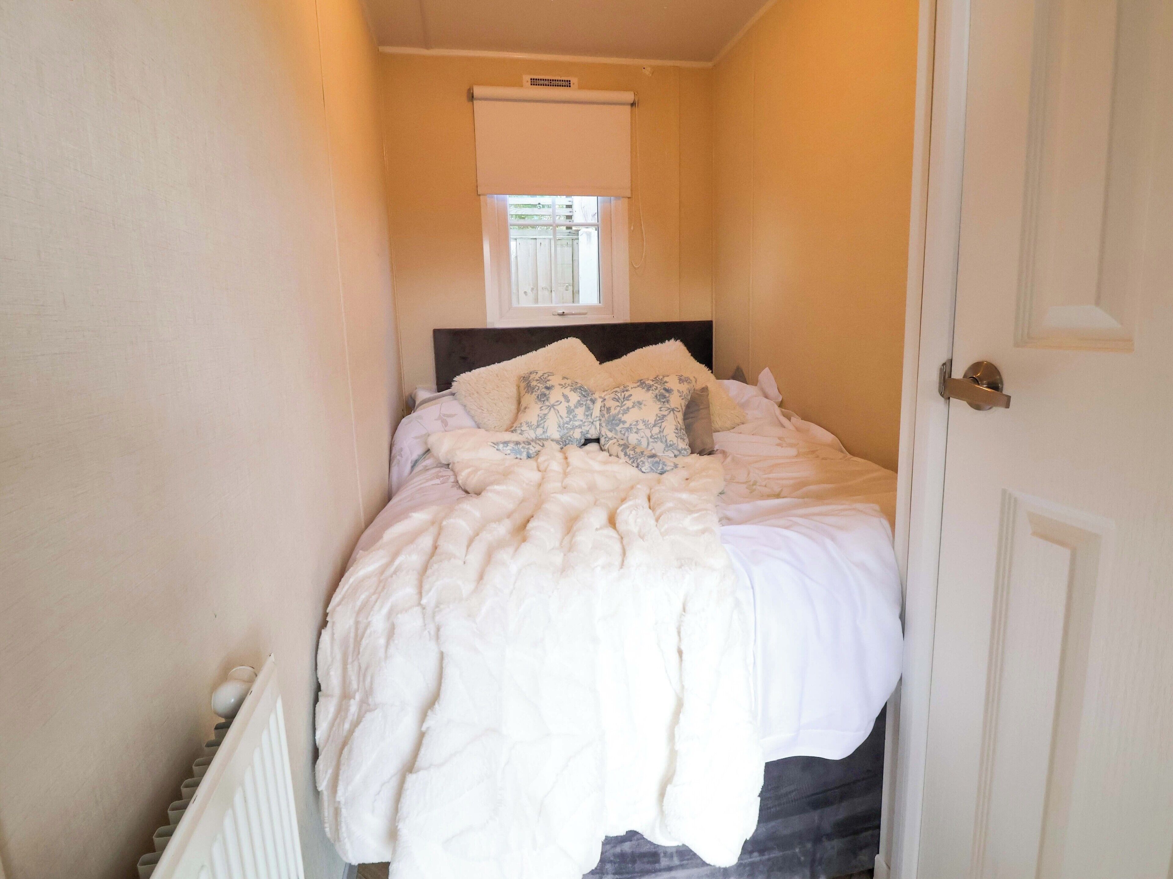 Una habitación estrecha y bien iluminada con una cama centrada frente a la pared, una ventana con persianas y una puerta a la derecha.