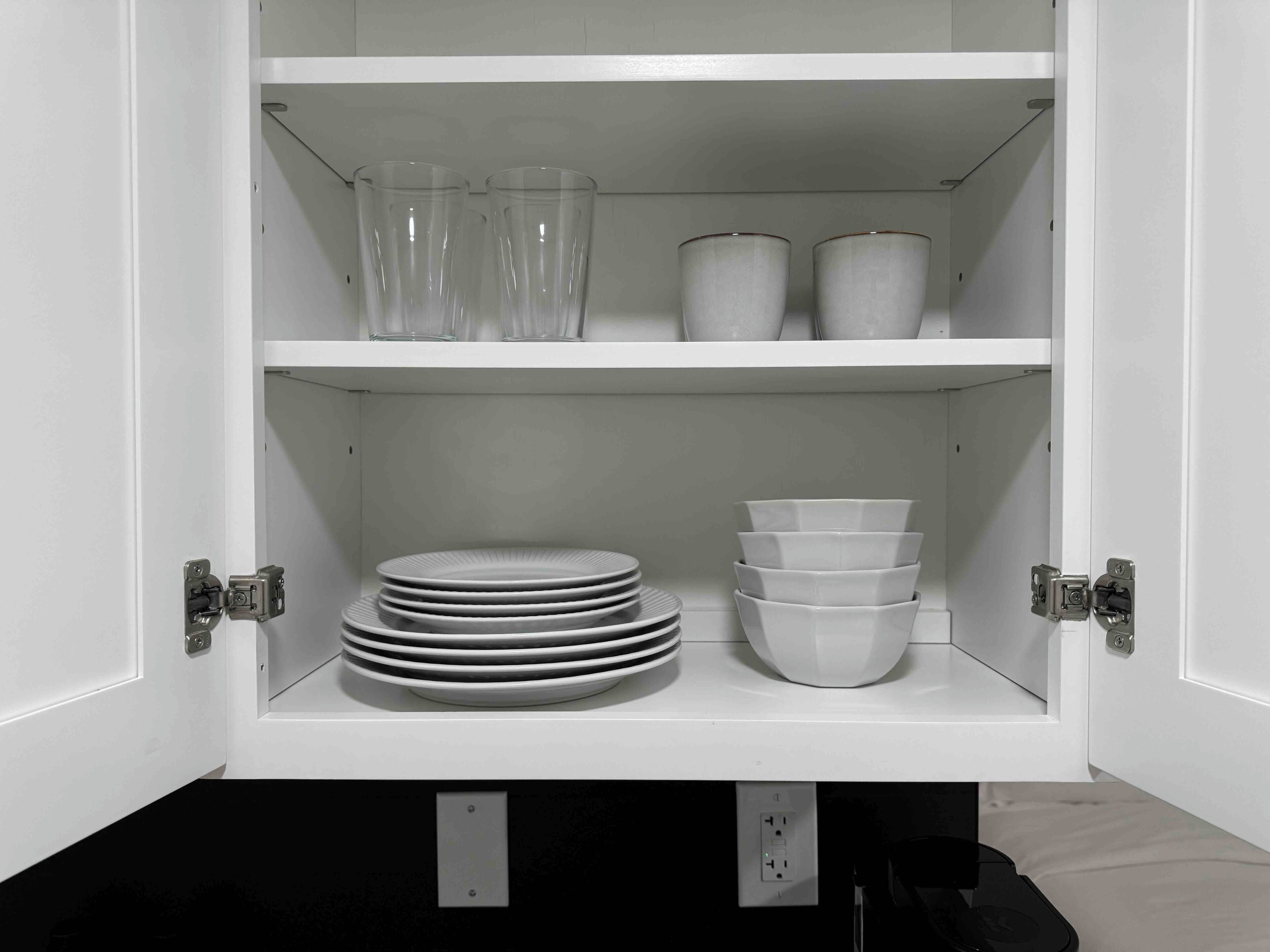 Un meuble blanc avec des étagères contenant des verres transparents, des assiettes blanches et des bols blancs.