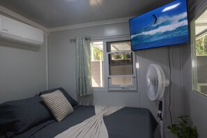 Une chambre à coucher avec un lit, un téléviseur fixé au mur, une fenêtre avec des rideaux et un ventilateur fixé au mur.
