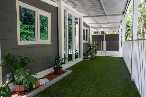Une véranda couverte avec un revêtement d’herbe artificielle, des plantes en pot et une vue sur la verdure environnante.