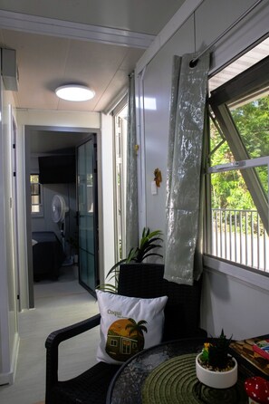 A compact living space with a glass door leading to a balcony, a small table with a plant, and a pillow with a tropical design. - Coquí Tiny House 1 – Modern & Cozy Retreat in Rincón (Rincón)