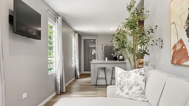 Un salon moderne avec un canapé, une table basse et un téléviseur fixé au mur. On aperçoit à l’arrière-plan un coin repas avec une table et des chaises.