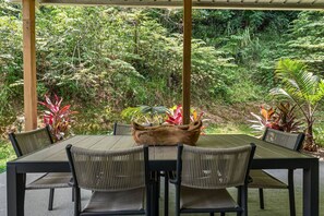 An outdoor dining area with a wooden table and chairs, surrounded by lush greenery and plants. - Vagones del Río | River Access | Sleeps 8 (Rincón)