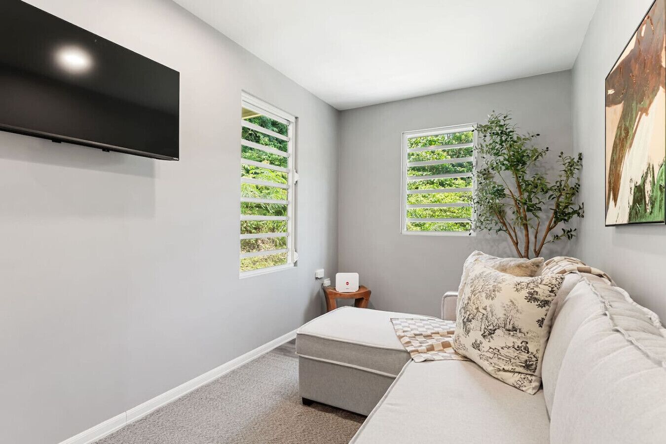 Un dormitorio moderno con un sofá gris, un televisor de pantalla plana montado en la pared, dos ventanas con persianas blancas, una mesita de noche con una lámpara y un cuadro en la pared.