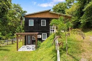A two-story black building with a wooden roof, surrounded by lush greenery and a white fence. - Vagones del Río | River Access | Sleeps 8 (Rincón)