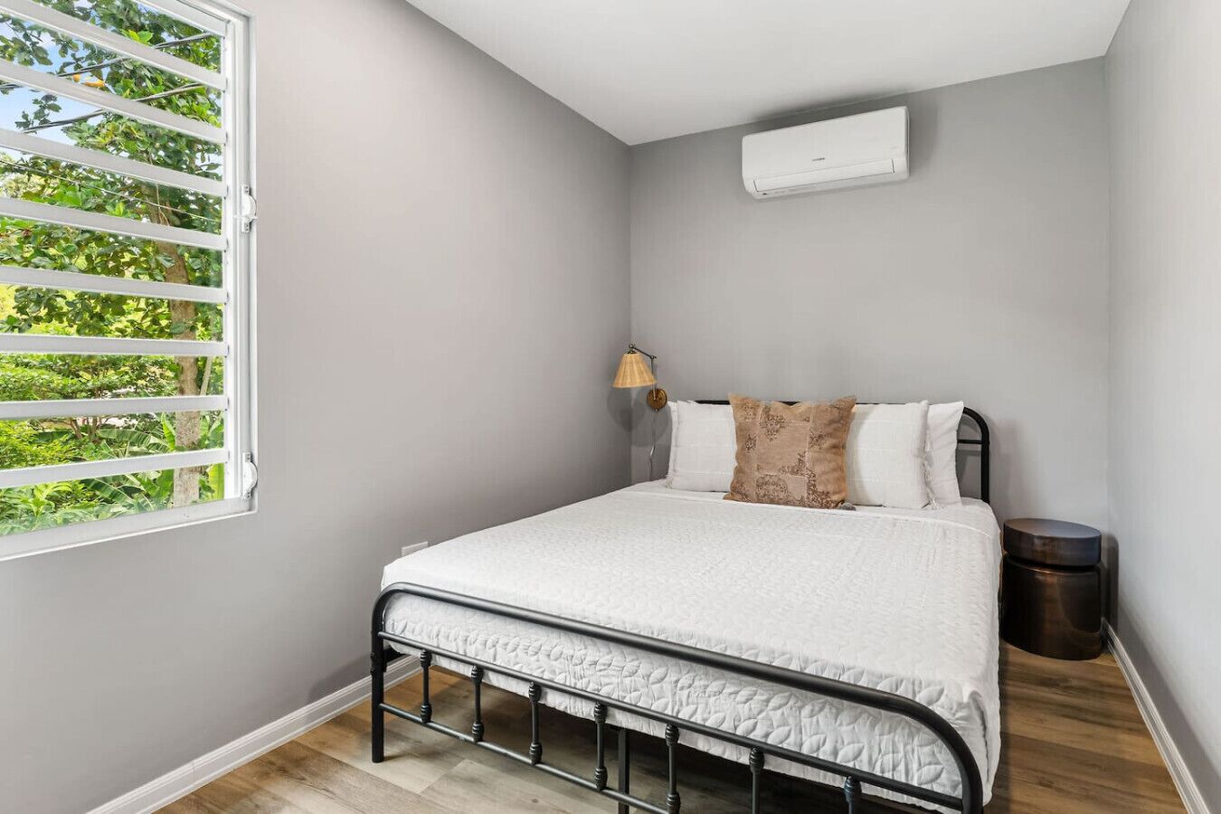 A bedroom with a bed, a window with blinds, a bedside lamp, and an air conditioning unit.