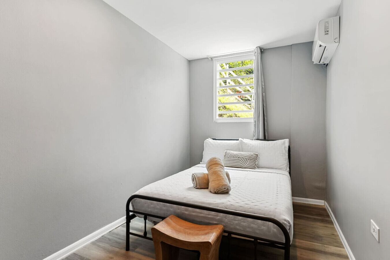 Une petite chambre à coucher, bien rangée, avec un lit simple, un tabouret en bois et une fenêtre avec des rideaux.