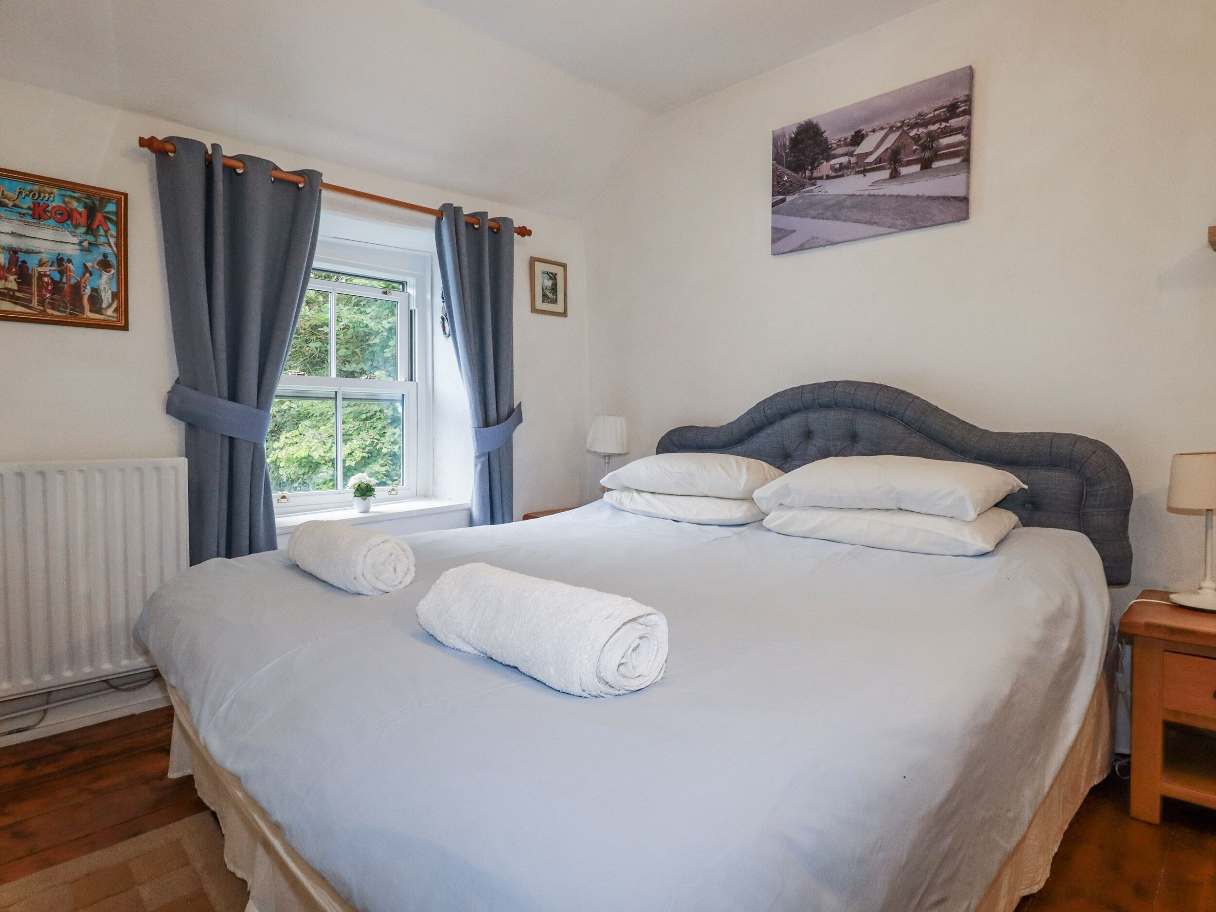 A neatly made bed with white linens and pillows, a window with curtains, a radiator, and a framed picture on the wall.