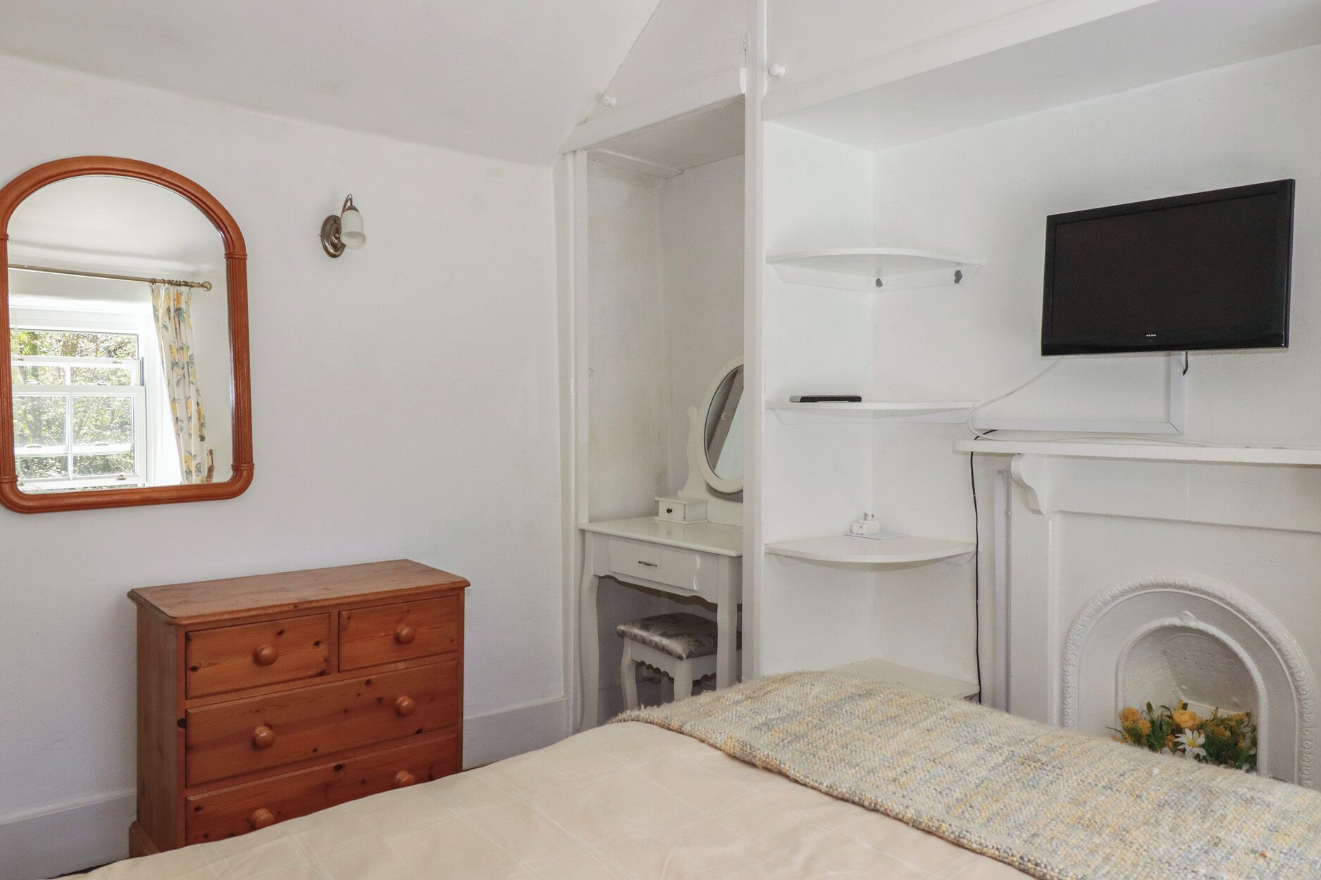 A bedroom with a bed, dresser, vanity, mirror, television, and fireplace.