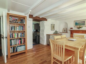 A dining area with a wooden table and chairs, a bookshelf with books, a kitchen in the background, and a framed picture on the wall. - Blowing House Cottage (Perranporth)