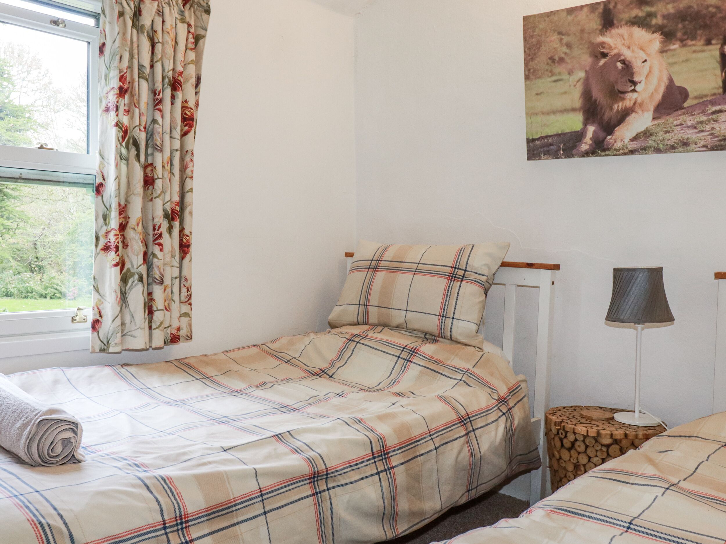 A bedroom with a plaid bedspread, a window with floral curtains, a wooden side table, and a lamp.