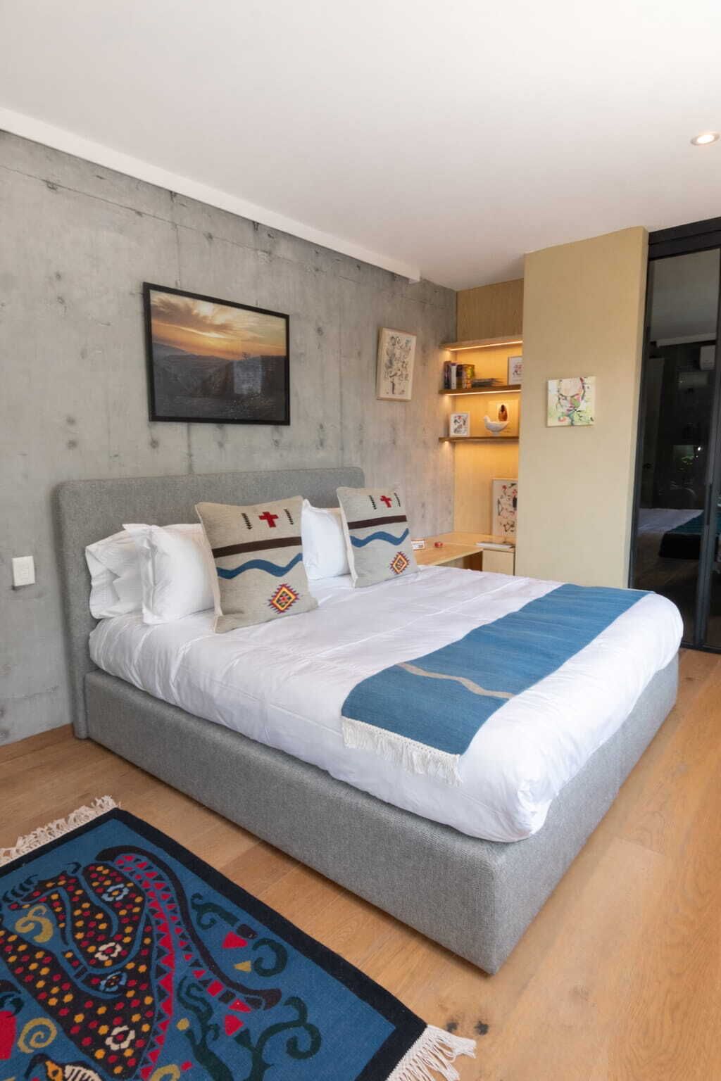 A modern bedroom with a grey bed, white bedding, and a blue throw blanket. There is a colorful rug on the floor and a painting on the wall.
