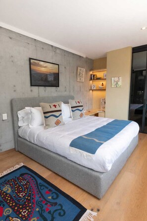 A modern bedroom with a grey bed, white bedding, and a blue throw blanket. There is a colorful rug on the floor and a painting on the wall. - Casa Gama Gabriel | Art & Design in Northern Rome (Ciudad de México)