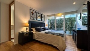 Ein modernes Schlafzimmer mit einem großen Bett, Nachttischen, einem Schreibtisch und Blick auf Grünpflanzen durch große Fenster.