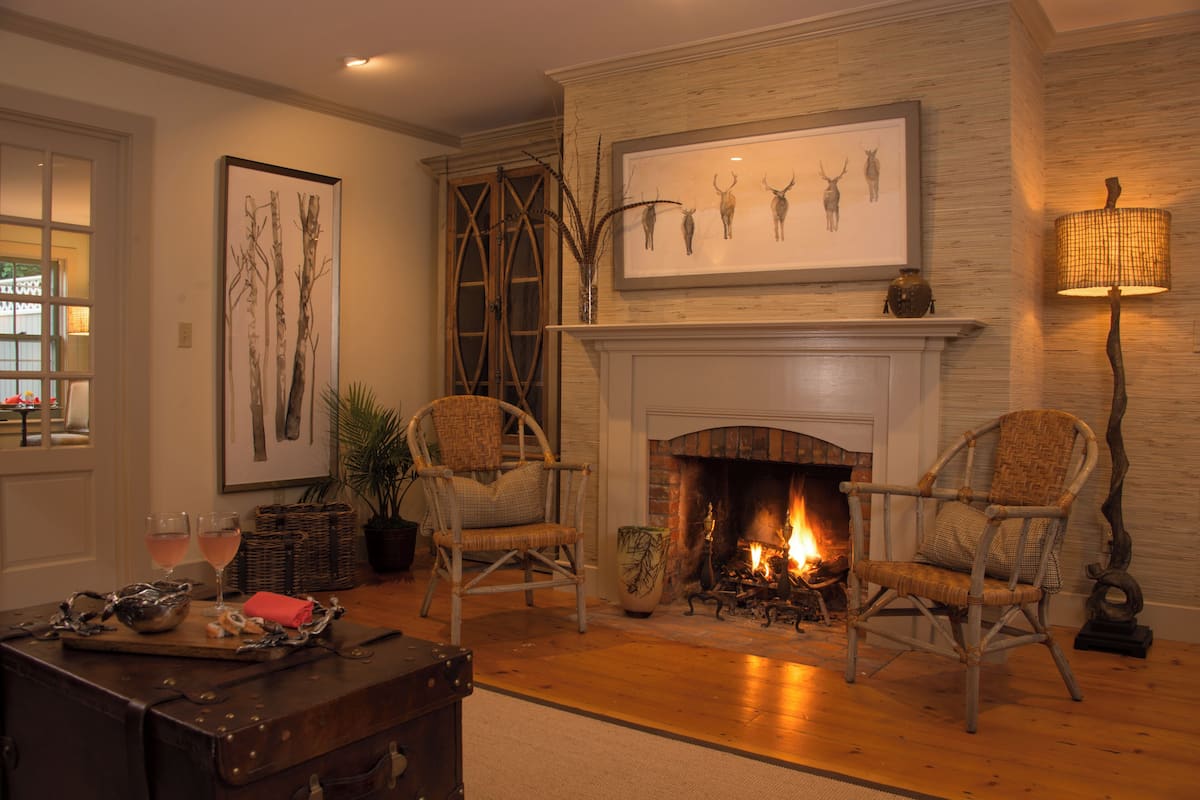 Una sala de estar acogedora con chimenea, dos sillones de mimbre, una mesa con bebidas y una obra de arte de un ciervo enmarcada sobre la repisa.