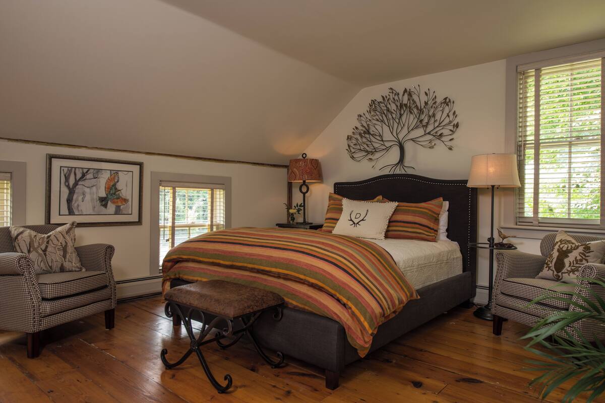 Un dormitorio con una cama grande, dos sillas, un banco, un cuadro de un árbol y dos lámparas.