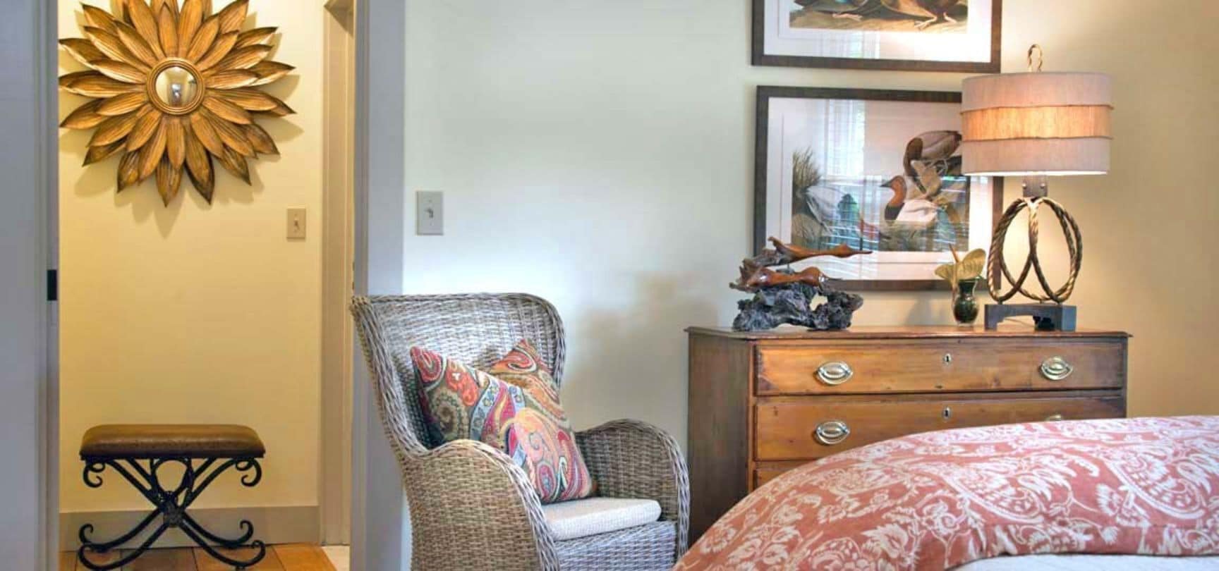 Une chambre à coucher avec un fauteuil en osier, une commode en bois, un miroir et un lit recouvert d’une couvre-lit à motifs.