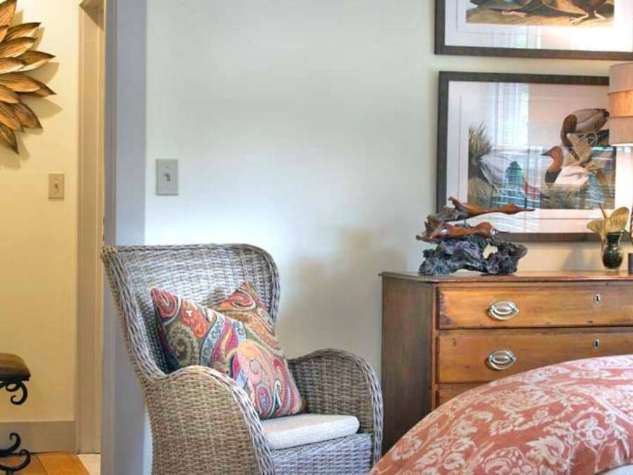 Une chambre à coucher avec un fauteuil en osier, une commode en bois, un miroir et un lit recouvert d’une couvre-lit à motifs.