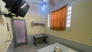 Una habitación de hotel pequeña y sencilla con una cama, un escritorio, un ventilador de techo, un televisor y una ventana con cortinas.