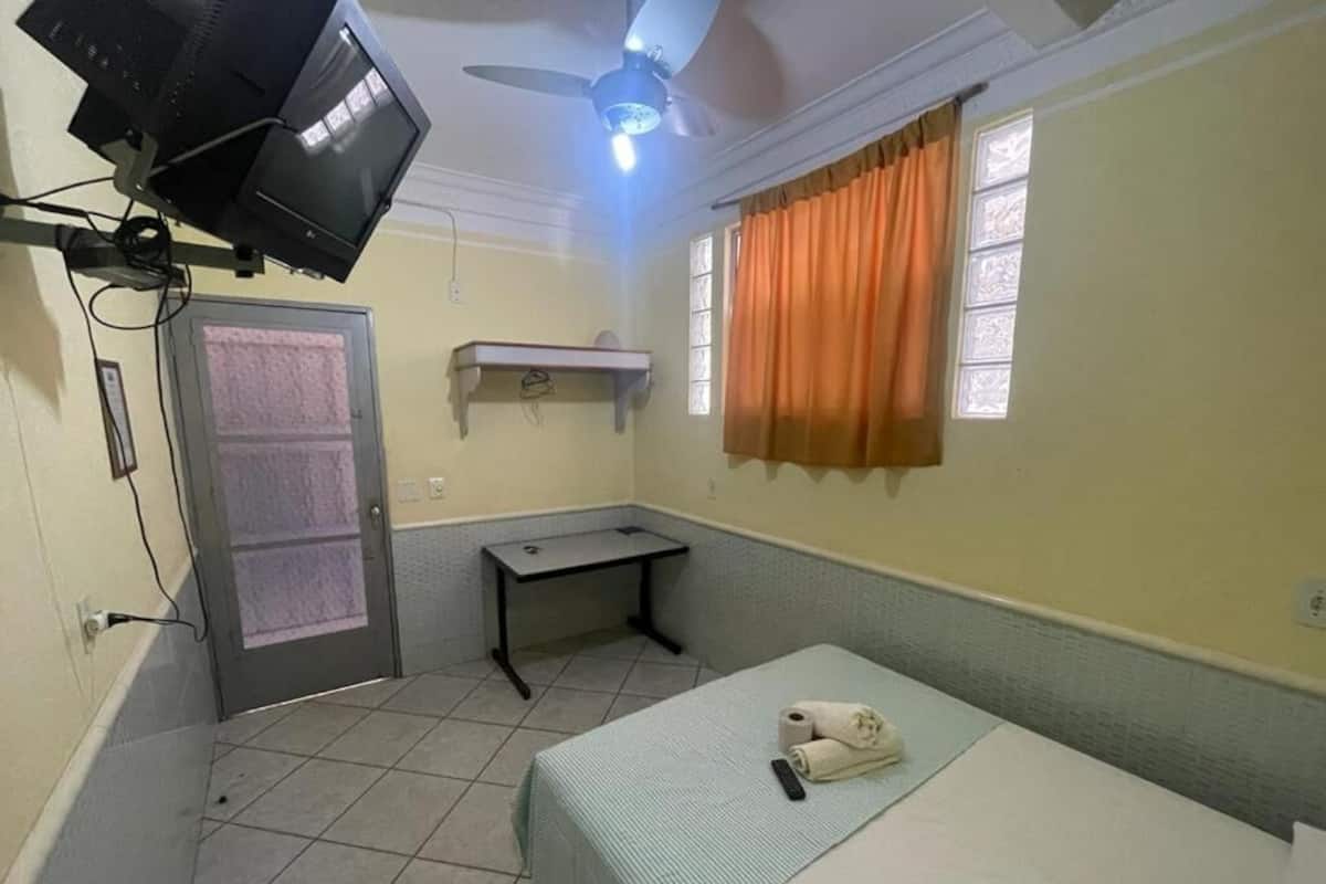 A small, single-room hotel room with a bed, a desk, a ceiling fan, a TV, and a window with curtains.