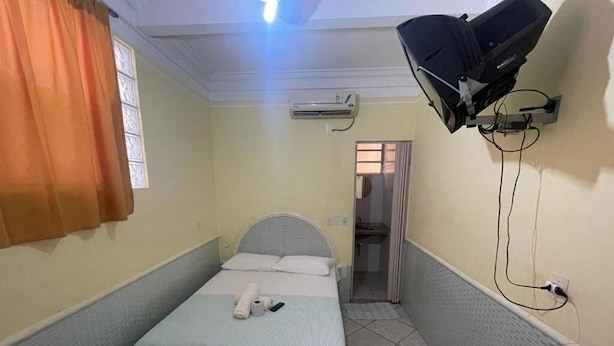 Una habitación de hotel pequeña y estrecha con una cama individual, un ventilador de techo, un equipo de aire acondicionado y un televisor montado en la pared.
